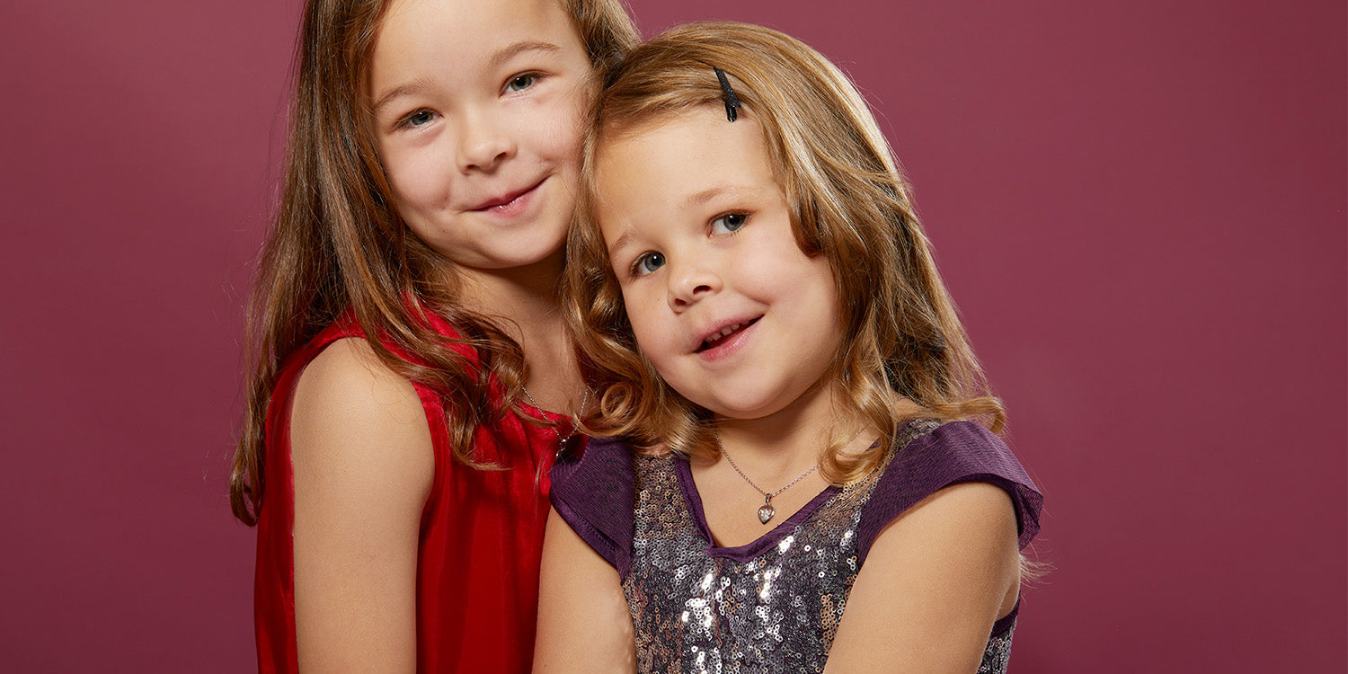 Zwei lächelnde Mädchen in festlichen Kleidern - eines trägt eine zarte silberne Halskette mit Anhänger