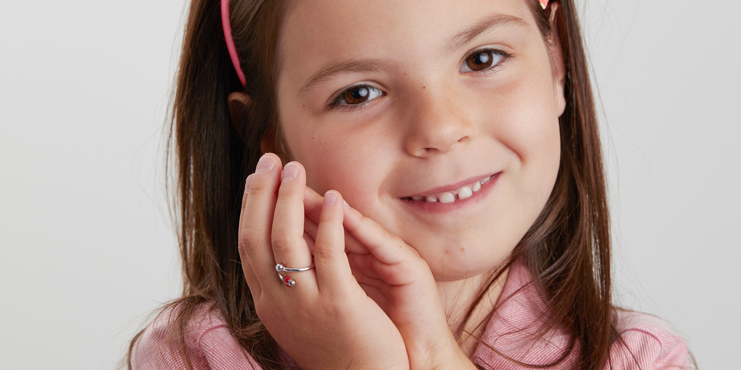 Lächelndes junges Mädchen mit braunen Haaren und rosa Haarreif trägt einen zierlichen Kinderring mit Marienkäfer-Motiv, Hand elegant zur Wange geführt, trägt rosa Oberteil vor weißem Hintergrund