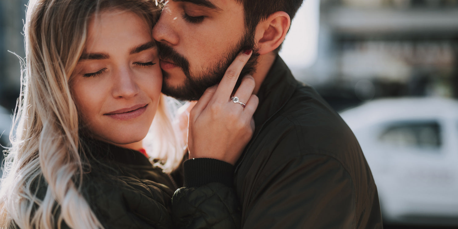 Verliebtes Paar in inniger Umarmung, die blonde Frau trägt einen eleganten Verlobungsring mit Diamant, während sie zärtlich das Gesicht ihres Partners berührt, romantische Szene im warmen Sonnenlicht