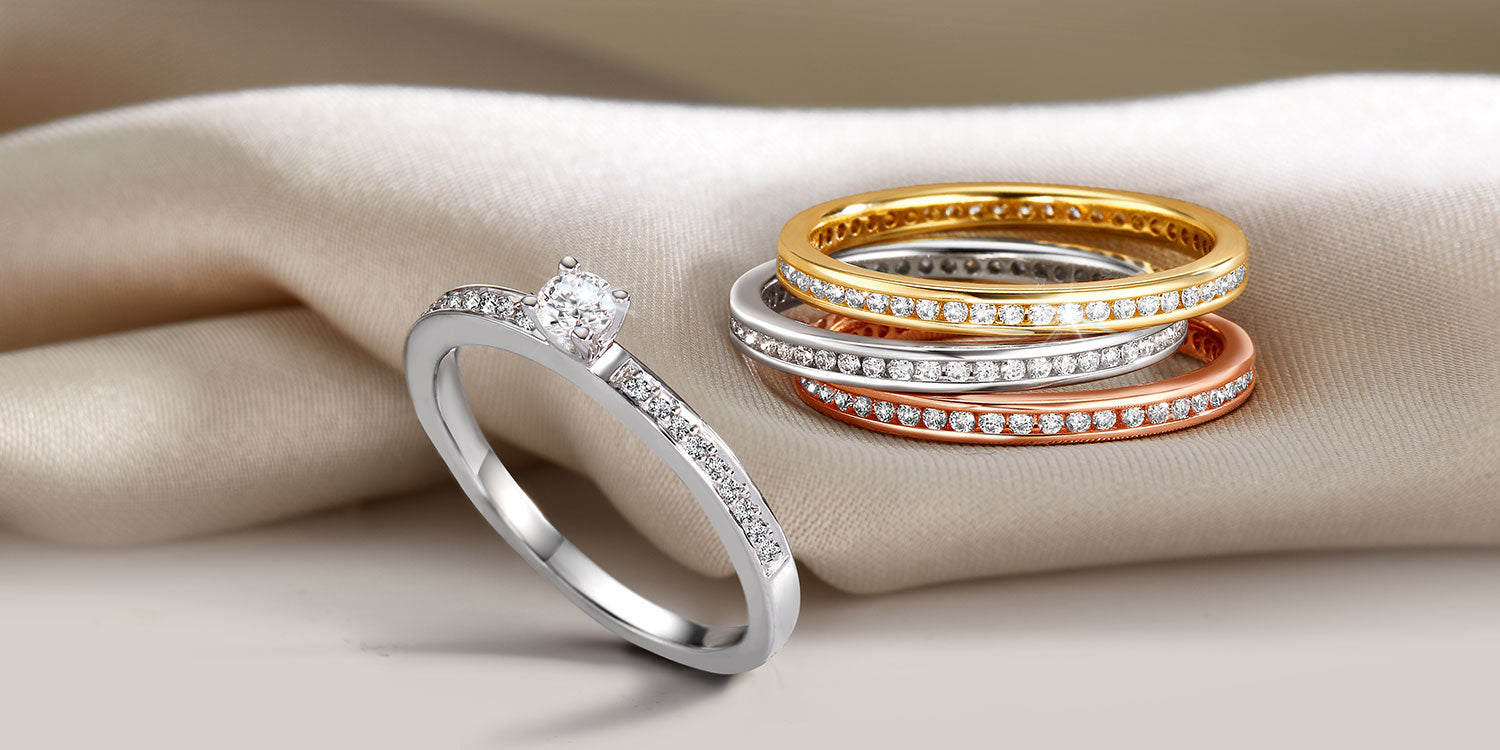 Eleganter Solitär-Verlobungsring aus Weißgold mit Diamant und Pavé-Fassung neben einem Set aus vier gestapelten Memory Ringen in Gold, Weißgold und Roségold mit Diamantbesatz, präsentiert auf cremefarbenem Seidenstoff